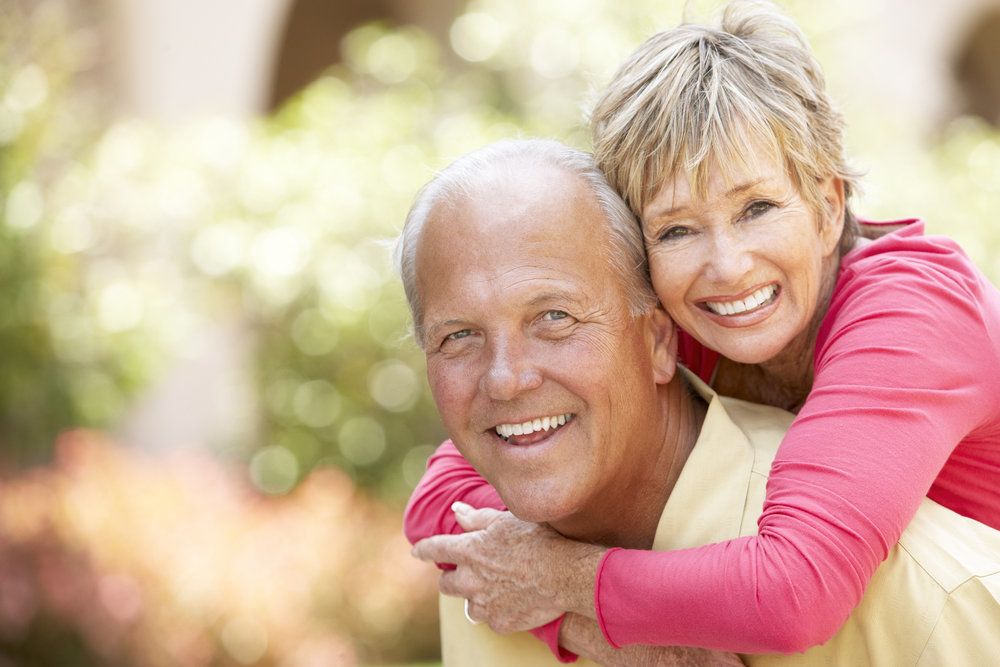 An elderly woman wearing a pink shirt grabs an elderly man in an embrace from his back.