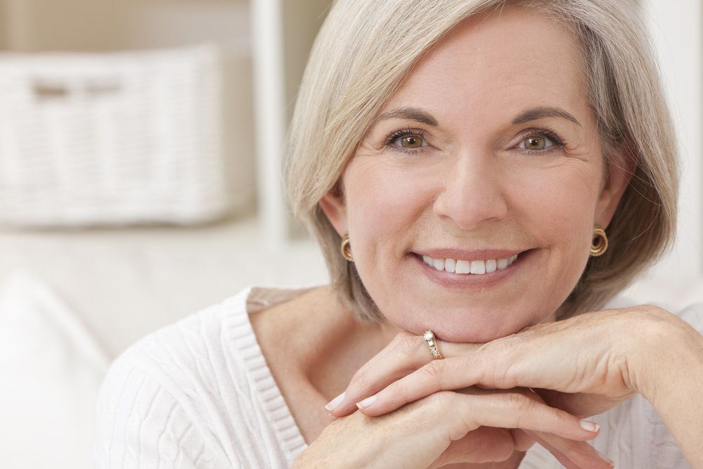 An elderly woman with a gray bob haircut smile happily with her hands held to her chin.